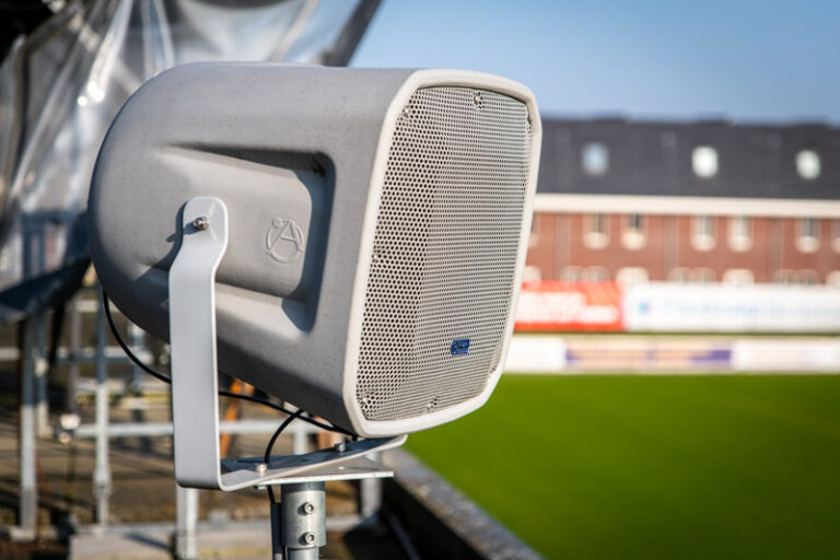Close up van AtlasIED speaker op het dak van een tribune, met op de achtergrond het groene voetbalveld.