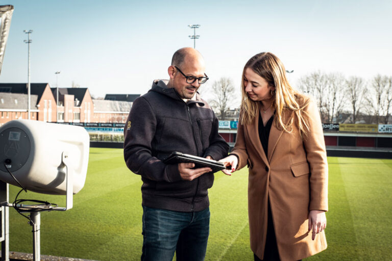 Man en vrouw op het dak van een voetbal tribune met een tablet en een speaker tijdens een geluidsdemonstratie, op de achtergrond het groene voetbalveld en een blauwe lucht.