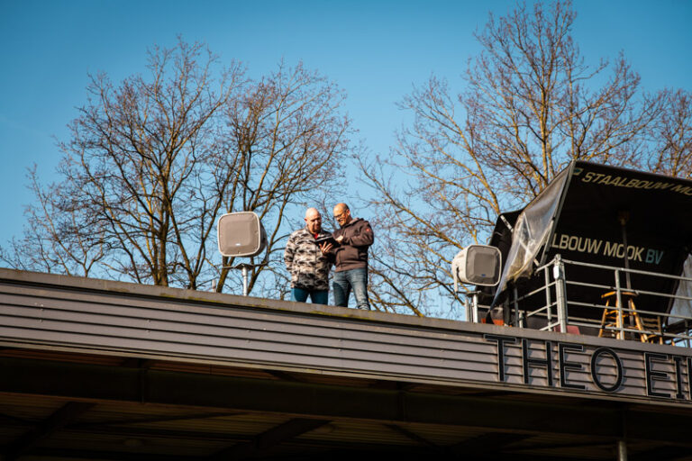Twee mannen met een tablet bovenop het dak van de tribune op een voetbalveld, tussen twee AtlasIED speakers in.