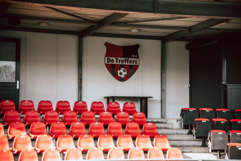 Rode stoeltjes van de tribune op het voetbalveld van De Treffers in Groesbeek, met op de achtergrond en clublogo.