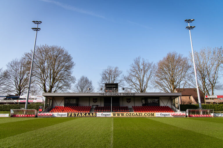 Tribune van de voetbalvereniging De Treffers, met op de voorgrond het groene voetbalveld en op de achtergrond een strakblauwe lucht.
