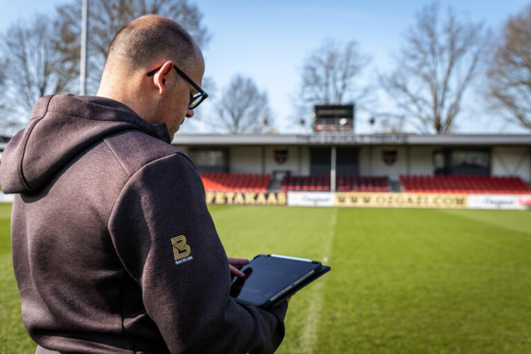 Man met tablet in het midden van een voetbalveld, met op de achtrond de tribune, het groene veld en een blauwe lucht.