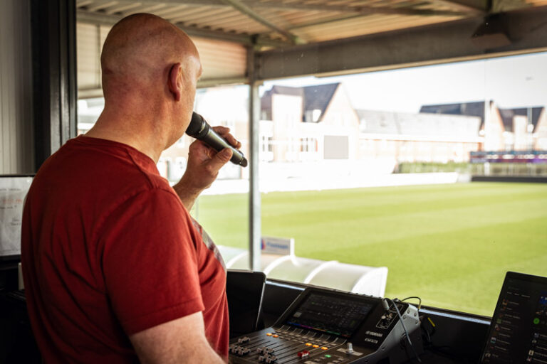 De live omroeper van De Treffers die met een microfoon aan zijn mond richting het groene voetbalveld kijkt.