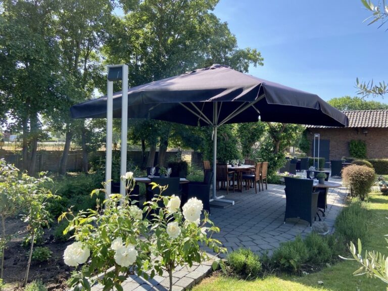 Het terras met open parasollen in de tuin van Kasteel Eyckholt. Er staat een zonnetje met een strakblauwe lucht en er is veel groen te zien.