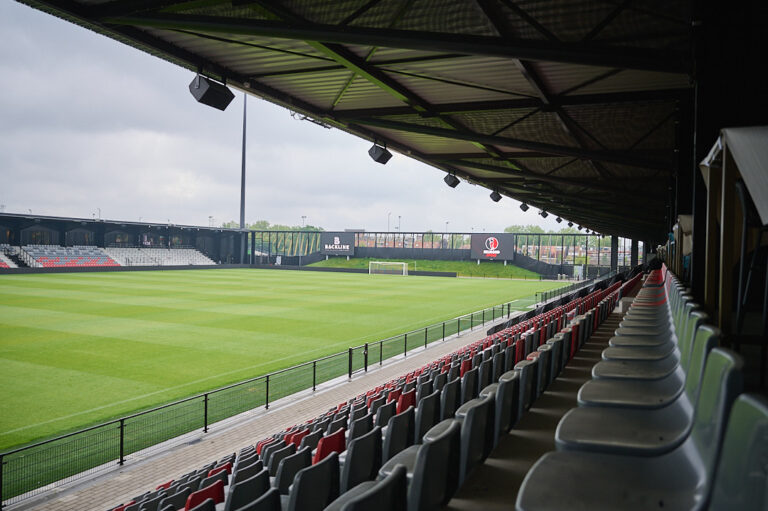 Doorkijk vanaf rechtertribune naar grote schermen aan het voetbalveld, met speakers op een rij aan overkapping.