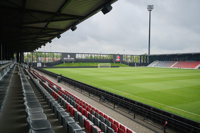 Doorkijk vanaf linkertribune naar grote schermen aan het voetbalveld, met speakers op een rij aan overkapping.