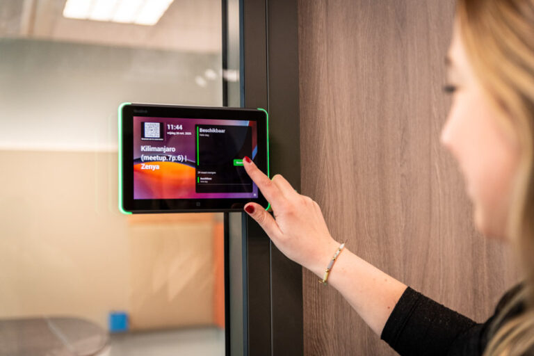 Vrouw die op de reserveer knop van het roombooking panel wil klikken, dat aan de buitenzijde van de vergaderzaal hangt.