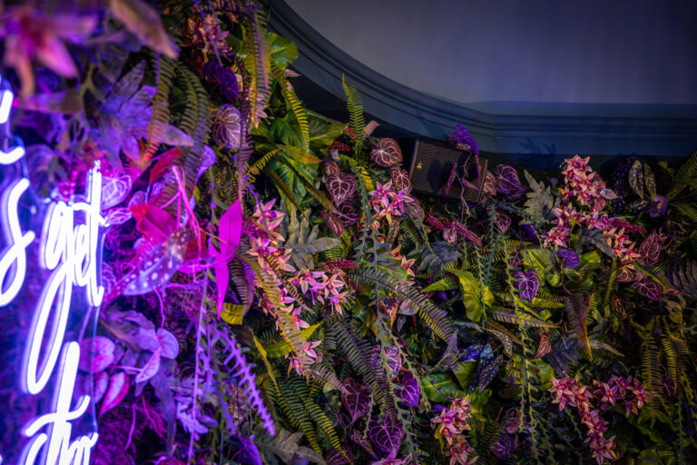 MAG audio speaker gemonteerd aan wand met kunstbeplanting en bloemen.